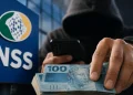 Pessoa encapuzada segurando dinheiro e celular em frente ao logotipo do INSS, representando golpe financeiro contra aposentados e pensionistas.